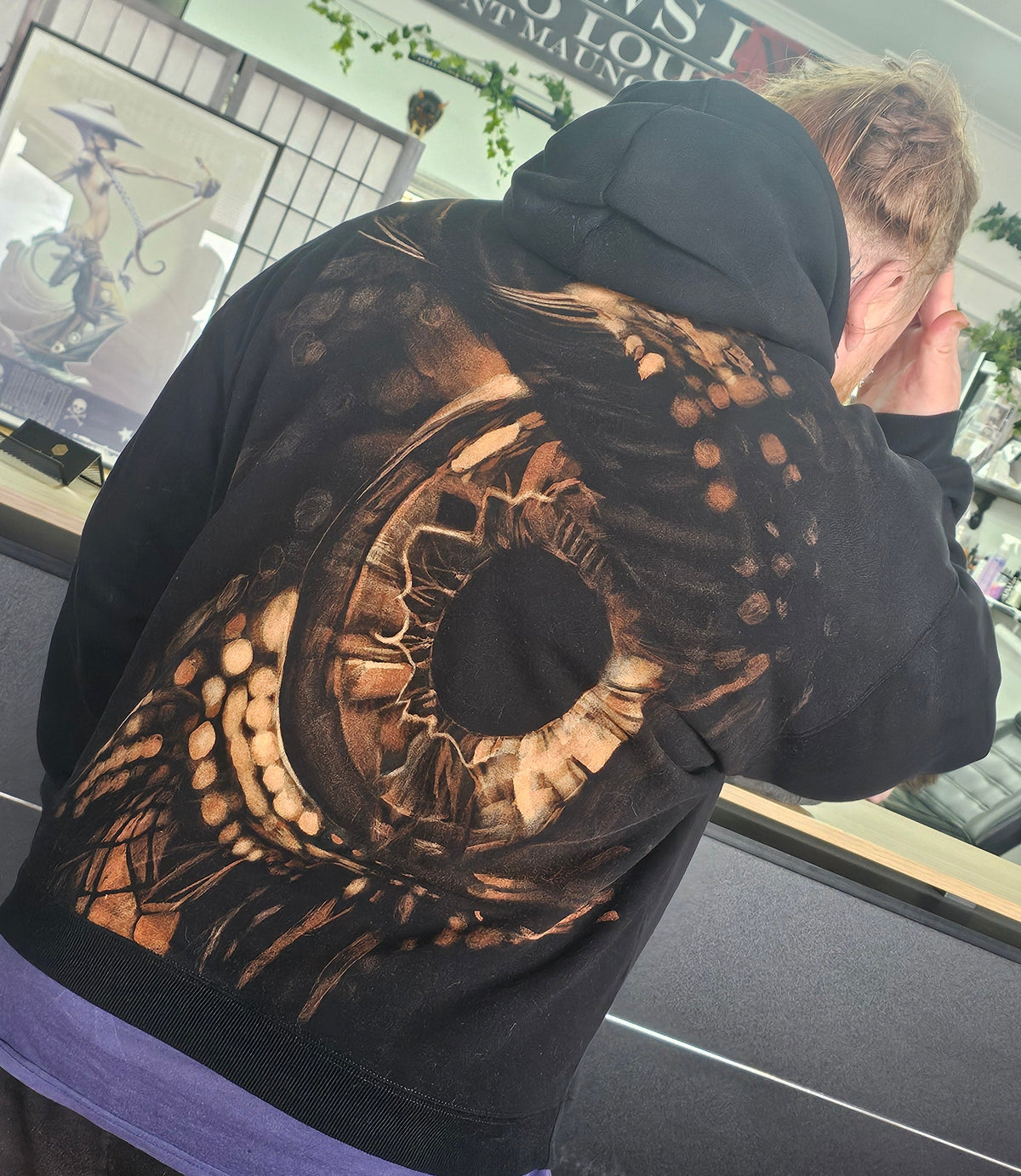 A person wearing a black hoodie with a large, detailed brown and beige graphic print on the back, standing inside a tattoo studio.